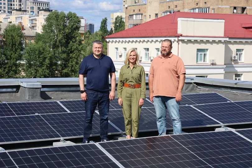 Троє людей стоять серед сонячних панелей на даху міської будівлі